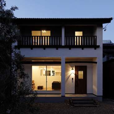 The image shows a modern two-story house at dusk, featuring large windows, a wooden balcony, a warm-lit cozy interior, and landscaped outdoor steps leading to the entrance.