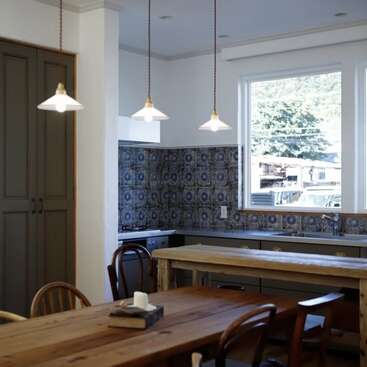 This cozy kitchen features patterned blue tiles, wooden furniture, three pendant lights, a large window with outside views, and a rustic, inviting dining table centerpiece.