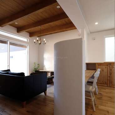 This cozy living space features exposed wooden ceiling beams, a chandelier, large windows, a black sectional sofa, a dining area with wooden furniture, and abundant natural light.