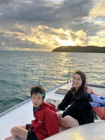 A boy and a woman sit on a boat at sunset, smiling and enjoying the scenic ocean view. Dramatic clouds and a bright sky background.