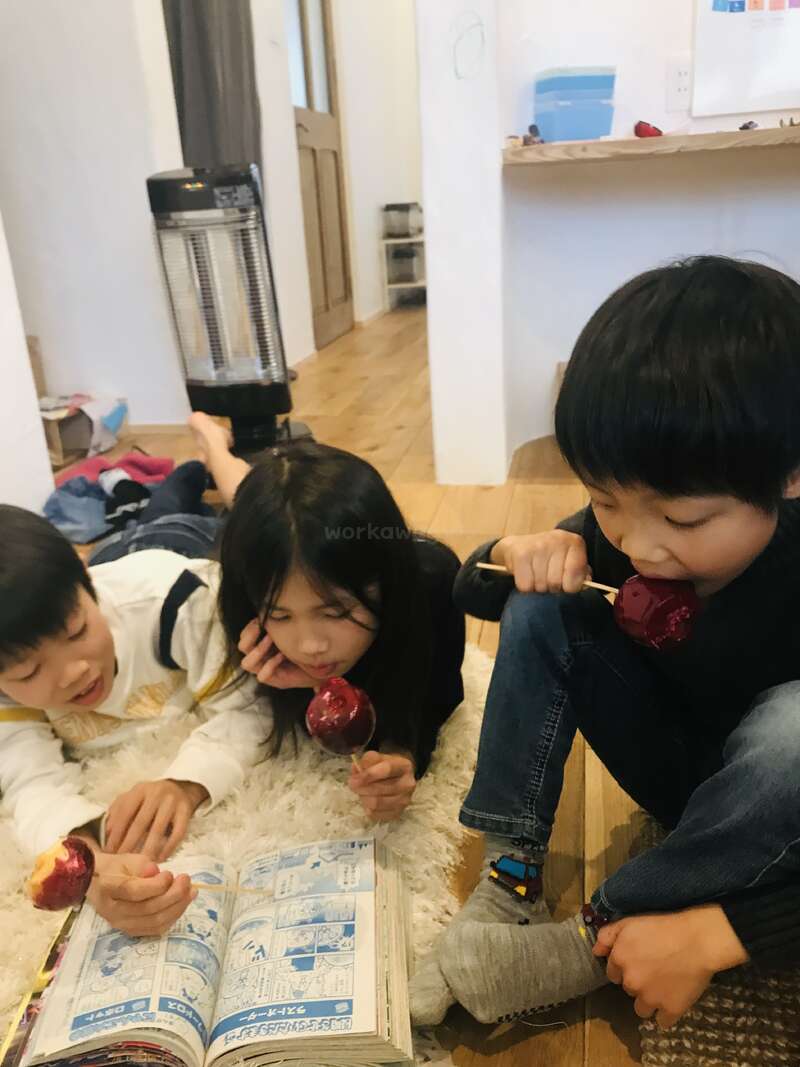 Three children sit together indoors, enjoying candied apples. Two are lying on a rug, reading a manga book, while the third sits nearby, happily snacking. Cozy atmosphere.