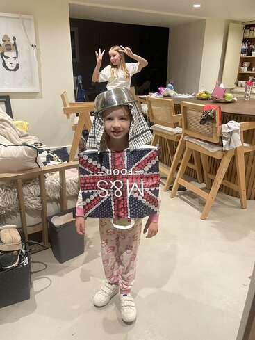 A young girl wears a pot on her head and a Union Jack box as armor. Another girl poses playfully behind her in a cozy kitchen.