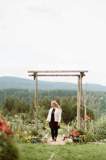 Eine Frau steht unter einem hölzernen Gartenbogen, umgeben von bunten Blumen und Grünpflanzen, mit malerischen bewaldeten Hügeln und einem bewölkten Himmel im Hintergrund. Friedliche Atmosphäre.