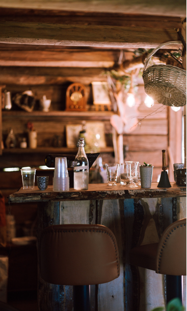 Eine rustikale Holzbar mit Hockern, Gläsern und einer Flasche, umgeben von gemütlicher Einrichtung, warmer Beleuchtung und Vintage-Artikeln, schafft eine einladende, heimelige Atmosphäre.