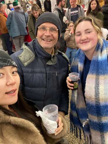 Drei Personen lächeln und halten Getränke bei einer geschäftigen Veranstaltung im Freien. Die Gruppe sieht fröhlich aus, trägt warme Kleidung und ist von anderen umgeben, die die festliche Atmosphäre genießen.