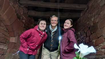 Drei Menschen lächeln und posieren zusammen in einem rustikalen Steinbau. Zwei Frauen tragen kastanienbraune Jacken, und ein Mann steht zwischen ihnen. Eine weiße Blume ist zu sehen.
