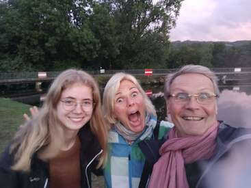 Drei Menschen posieren für ein fröhliches Selfie im Freien in der Nähe eines Flusses, einer Brücke und von Bäumen. Sie lächeln und machen lustige Grimassen und genießen einen lebhaften Moment zusammen.