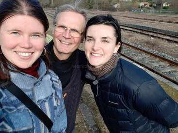Drei Personen lächeln und machen ein Selfie im Freien in der Nähe von Bahngleisen. Sie tragen Jacken, und das Wetter scheint kalt und sonnig zu sein. Im Hintergrund sind entfernte Häuser zu sehen.
