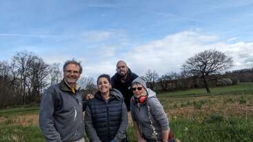 Vier Personen posieren lächelnd und lässig gekleidet auf einer Wiese. Hinter ihnen stehen Bäume und ein blauer Himmel mit vereinzelten Wolken, die eine entspannte Atmosphäre schaffen.