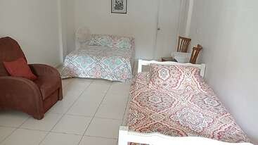 Esta imagem mostra um quarto arrumado com uma cama de casal estampada, um sofá-cama estampado, uma poltrona marrom, um ventilador, duas cadeiras e um piso de azulejos brancos.