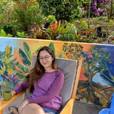 La imagen muestra a una mujer sentada en una silla de madera con una pared pintada y exuberantes plantas de fondo, que desprenden un ambiente sereno y natural.