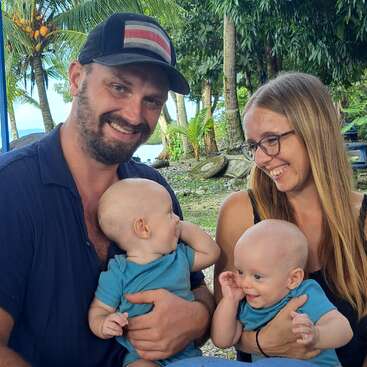 Un couple heureux est assis en plein air, chacun tenant un bébé souriant. Entourée d'une végétation tropicale luxuriante, la famille partage joie et rire dans ce moment d'amour.