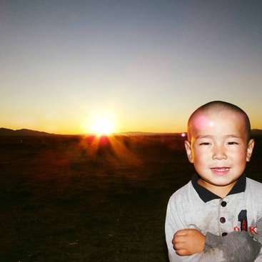 Das Bild zeigt einen Jungen, der vor einem atemberaubenden Sonnenuntergang steht. Die Sonnenstrahlen werfen ein warmes Licht auf sein Gesicht und die umliegende Landschaft.