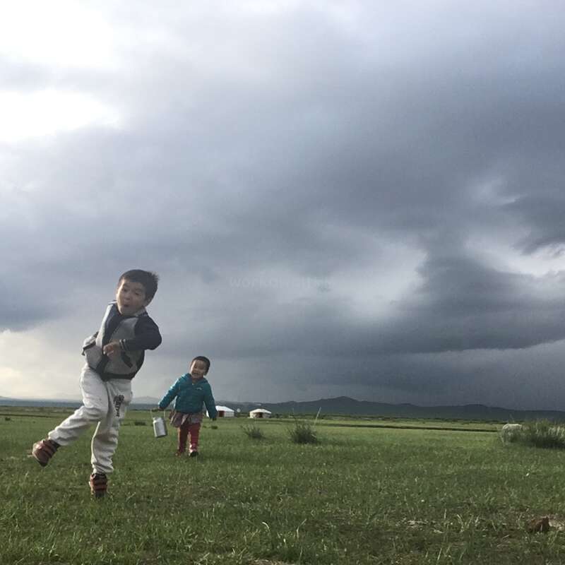 Zwei Kinder rennen unter einem stürmischen Himmel über eine grasbewachsene Wiese, während in der Ferne Pferde grasen und im Hintergrund eine Jurte zu sehen ist.