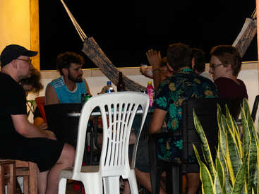 L'image représente un groupe de personnes assises autour d'une table sur un balcon la nuit, avec un hamac et des plantes visibles au premier plan.