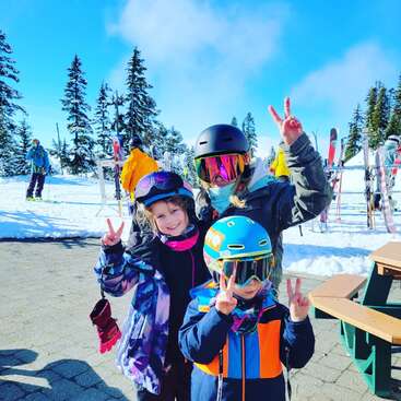 Das Bild zeigt eine Frau und zwei Kinder, die für ein Foto in einem Skigebiet posieren und beide mit ihren Händen ein Friedenszeichen machen. Sie tragen alle eine Winterausrüstung.
