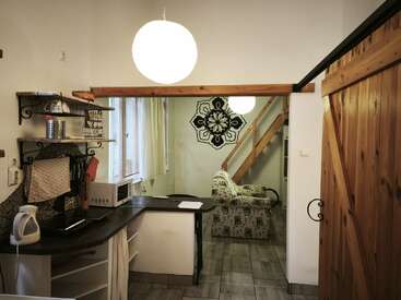This cozy studio features a small kitchen area, patterned armchair, decorative wall art, wooden barn door, staircase, large round light, and rustic shelves with kitchen essentials.