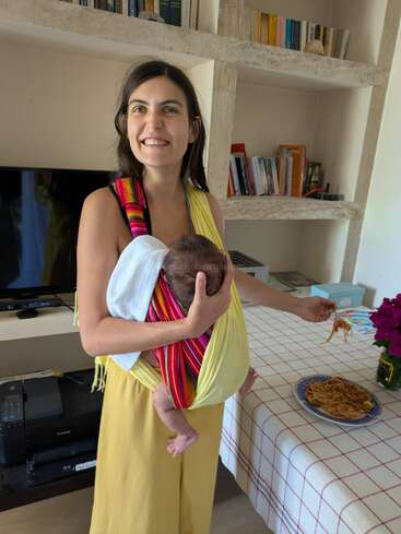 A smiling woman in yellow holds a baby in a colorful sling. She stands by a table with a plate of spaghetti, grabbing some food.