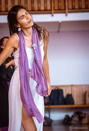 A woman in a white dress and purple scarf dances with eyes closed, expressing emotion. The background is indoors with bags, wooden bench, and soft lighting.