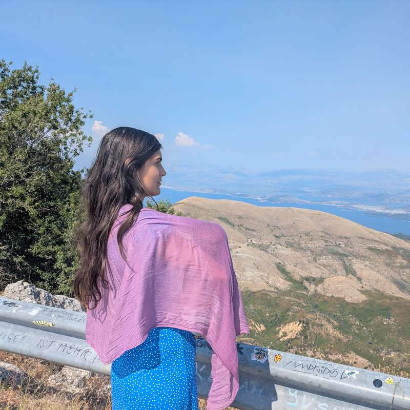 A woman with long brown hair, wearing a blue dress and purple shawl, stands by a guardrail, gazing at rolling hills and serene blue water.