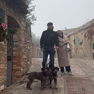 An older couple stands affectionately together, walking two curly-haired dogs on a cobblestone street adorned with murals and flowers, on a misty, peaceful day.