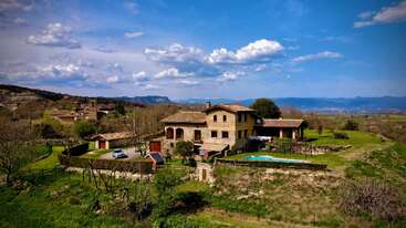 Eine rustikale Landvilla, umgeben von üppigem Grün, Gärten und Bäumen. Ein kleiner Pool und eine Terrasse laden zum Entspannen ein. Berge und blauer Himmel bilden eine friedliche Kulisse.
