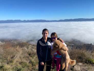 Eine Frau und ein Mädchen mit einem flauschigen Hund posieren auf einem grasbewachsenen Hügel über einem Meer aus Wolken, im Hintergrund sind unter strahlend blauem Himmel Berge zu sehen.