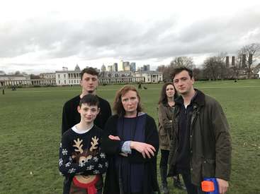 The image depicts a family of five standing on a grassy field, with a city skyline and large white building in the background, set against an overcast sky.