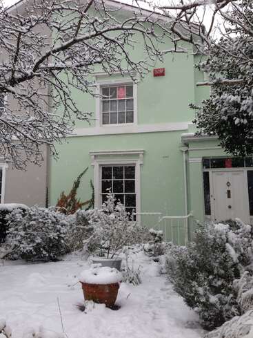 The image depicts a two-story house with a light green exterior, white trim, and a red sign on the front, surrounded by snow-covered bushes and trees.