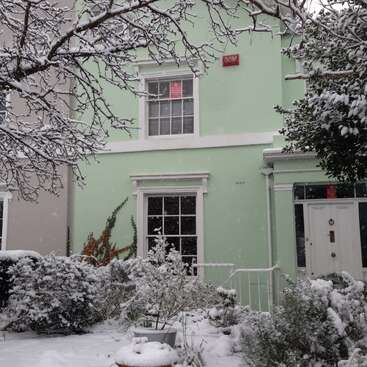 The image depicts a two-story house with a light green exterior, white trim, and a red sign, surrounded by snow-covered trees and bushes in the front yard.