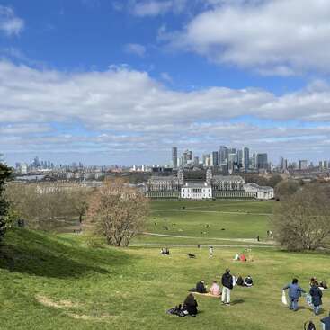 Die Leute entspannen sich und genießen einen sonnigen Tag auf einem grasbewachsenen Hügel, von wo aus sie unter einem teilweise bewölkten blauen Himmel auf historische Gebäude und die Skyline einer modernen Stadt blicken.