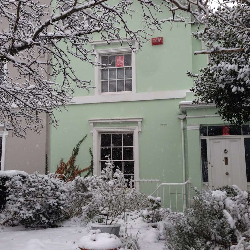The image depicts a two-story house with a light green exterior, white trim, and a red sign, surrounded by snow-covered trees and bushes in the front yard.
