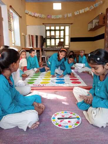 Um grupo de crianças com uniformes azuis está jogando Twister dentro de casa, sentadas em um tapete. A sala está decorada com guirlandas de papel coloridas e tem luz do sol.