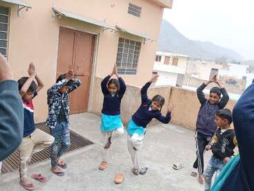 Um grupo de crianças está do lado de fora, fazendo uma pose de ioga, provavelmente a pose da árvore, com as mãos juntas acima da cabeça e equilibrando-se em uma perna.