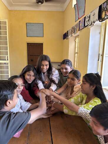 Um grupo de crianças sorridentes sentadas em volta de uma mesa de madeira, juntando as mãos no meio com entusiasmo, simbolizando união, amizade e trabalho em equipe numa sala de aula.