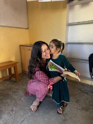 Uma mulher feliz se agacha enquanto uma menina beija sua bochecha. A menina segura um caderno, e as duas compartilham um momento feliz e emocionante dentro de uma sala de aula.
