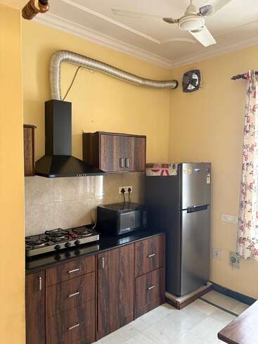 Essa imagem mostra uma cozinha compacta com armários de madeira, fogão a gás, chaminé, geladeira, micro-ondas, paredes amarelas, ventilador de teto e cortinas florais na janela.