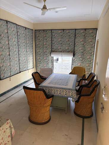 Uma sala aconchegante com cadeiras de vime ao redor de uma mesa coberta por uma toalha de mesa com estampa azul. Cortinas florais cobrem as janelas e um ventilador de teto está pendurado acima.