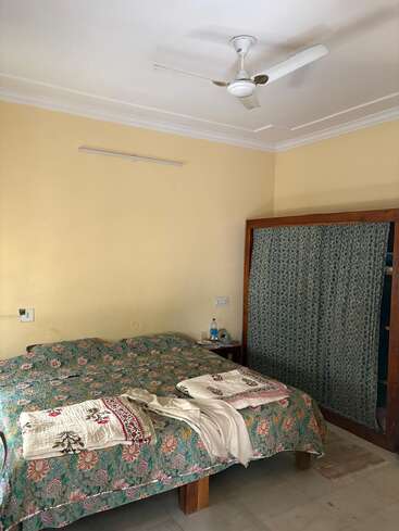 Um quarto aconchegante com colcha floral, toalha e xale colocados na cama, paredes amarelas, ventilador de teto, mesinha lateral e guarda-roupa com cortinas.