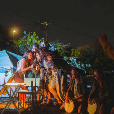 Um grupo de pessoas amontoadas canta em um microfone sob o céu noturno, iluminado por luzes quentes. Duas pessoas próximas tocam bateria, criando uma atmosfera animada.