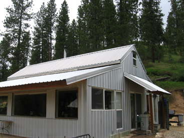 A imagem mostra uma pequena casa de um andar com telhado e revestimento de metal branco, situada em uma área arborizada com árvores altas ao fundo.