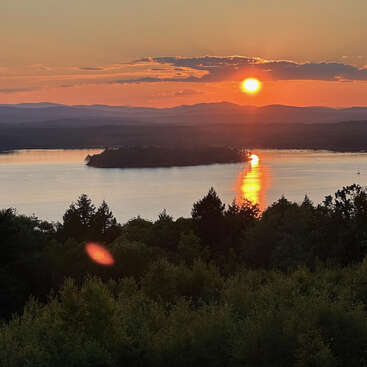 Ein krasser Sonnenuntergang taucht einen ruhigen See in orangefarbenes Licht, das sich wunderschön spiegelt, während die Silhouetten der Bäume und die fernen Berge eine friedliche, ruhige Naturlandschaft schaffen.