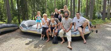 A group of nine people, including adults and children, sits on an inflatable boat in a wooded area, smiling and enjoying a sunny day outdoors together.