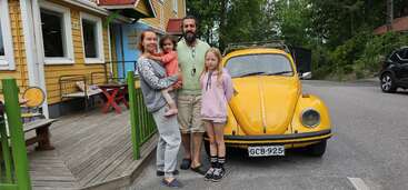 A family of four stands together outdoors beside a classic yellow Volkswagen Beetle. They are smiling, with a yellow wooden building and green trees behind them.