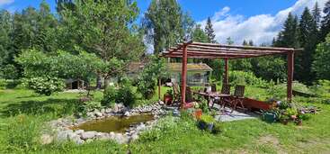 A peaceful garden scene with a wooden pergola, outdoor seating, a small pond surrounded by stones, lush greenery, trees, and a blue sky above.