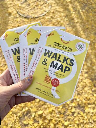 Eine Hand hält mehrere "Walks & Map"-Prospekte für Hida, die für ein glutenfreies Projekt werben. Im Hintergrund sind gelbe, gefallene Blätter zu sehen, die den Boden bedecken.