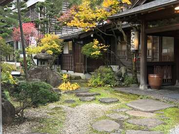Ein friedlicher japanischer Garten mit Steinlaternen, Trittsteinen, üppigem Grün und herbstlich gefärbten Bäumen umgibt ein traditionelles Holzgebäude, das Ruhe und kulturelle Schönheit ausstrahlt.