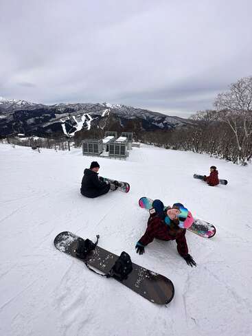 Drei Snowboarder ruhen sich auf einem verschneiten Berghang aus, umgeben von kahlen Bäumen und fernen schneebedeckten Gipfeln. Der Himmel ist bewölkt und schafft eine ruhige, winterliche Atmosphäre.