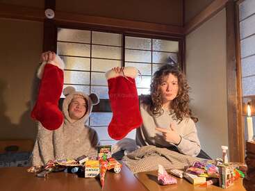 Zwei Menschen sitzen an einem Holztisch, der mit Snacks gedeckt ist. Beide halten rote Weihnachtsstrümpfe hoch, einer trägt ein Bärenkostüm, der andere zeigt auf den Strumpf.