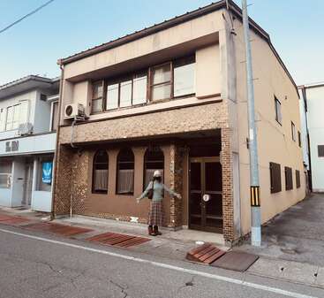 Eine Person steht vor einem zweistöckigen beigen Gebäude mit braunen Backsteinakzenten, großen Fenstern und einer Tür. Die Straße ist leer und friedlich.
